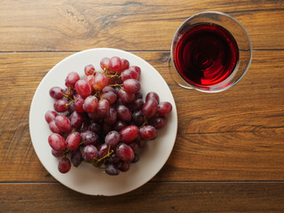 Fresh red grapes on a white plate and a glass of a red wine on a wooden table.