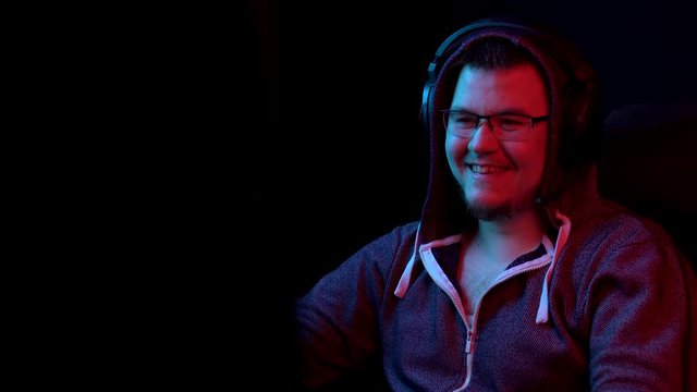 A young man sits at a computer with headphones. A man in a hood looks at a computer monitor and laughs. The camera zooms out from behind the monitor.