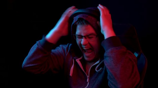 A young man sits at a computer and screams. The man in the hood looks at the monitor and curses with anger. The camera zooms out from behind the monitor