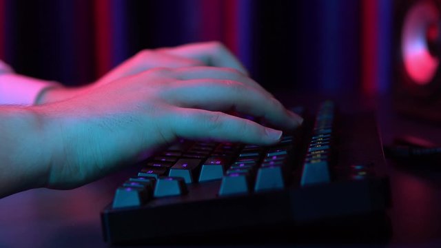 A young man is typing on a computer keyboard. Hands close up. Blue and red light falls on the hands.