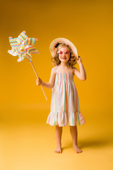 child girl blonde smiling in sunglasses and summer sundress holding a windmill stands on a yellow background. happy child. space for text