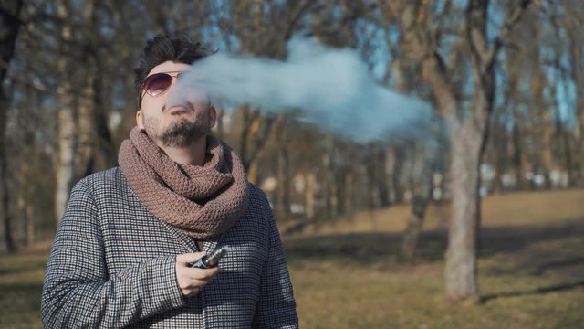 A young handsome bearded hipster man vaping an electronic cigarette in the park in the spring. Close up.