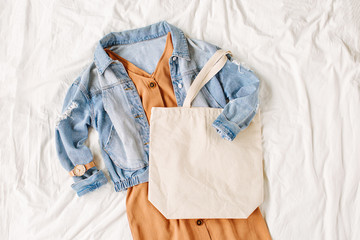 Blue jean jacket and  beige dress with tote bag on white bed. Women's stylish autumn outfit. Trendy clothes with white eco bag mockup. Flat lay, top view.
