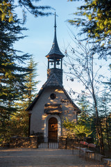 Fototapeta na wymiar Germanic small wooden church with two bells at the tower in La Cumbrecita, with a dirt road and a bench at the front of it, surrounded by evergreen trees