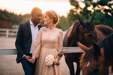 Happy smiling newlyweds standing on ranch surrounded by horses at sunset - obrazy, fototapety, plakaty