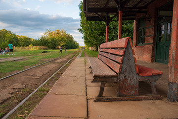 A wooden bench in an old abandoned train station with a view of the railroad with grass aroun it © LautaroFederico