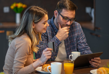 Happy couple using tablet and having breakfast in the kitchen