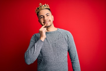 Young handsome man wearing golden crown of prince over isolated red background looking confident at...