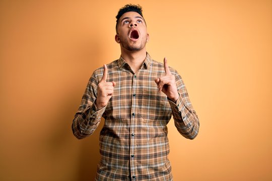 Young handsome man wearing casual shirt standing over isolated yellow background amazed and surprised looking up and pointing with fingers and raised arms.