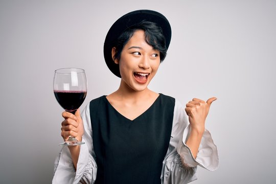 Young beautiful asian sommelier girl drinking glass of red wine over isolated white background pointing and showing with thumb up to the side with happy face smiling