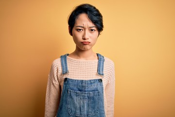 Young beautiful asian girl wearing casual denim overalls over isolated yellow background depressed and worry for distress, crying angry and afraid. Sad expression.