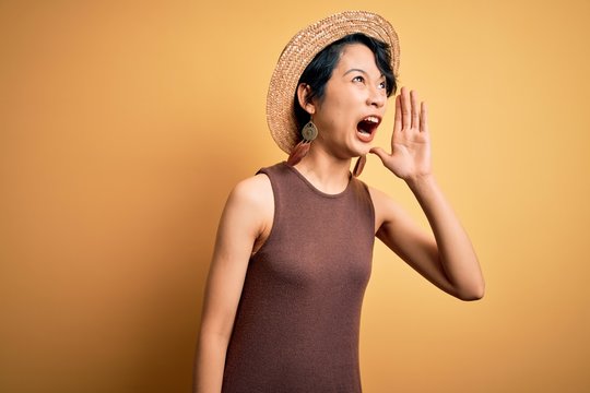 Young beautiful asian girl wearing casual t-shirt and hat over isolated yellow background shouting and screaming loud to side with hand on mouth. Communication concept.