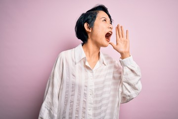 Young beautiful asian girl wearing casual shirt standing over isolated pink background shouting and screaming loud to side with hand on mouth. Communication concept.