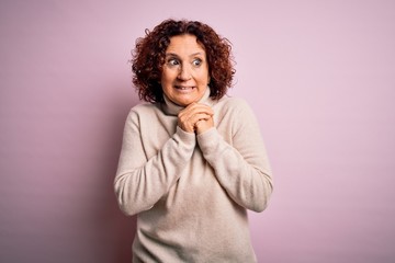 Middle age beautiful curly hair woman wearing casual turtleneck sweater over pink background laughing nervous and excited with hands on chin looking to the side