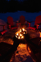 Cozy/romantic evening scene of warming bonfire on snow in winter with red chairs around. Canadian symbols of bear, paw and tree on the fire container. Banff National Park, Alberta, Canada