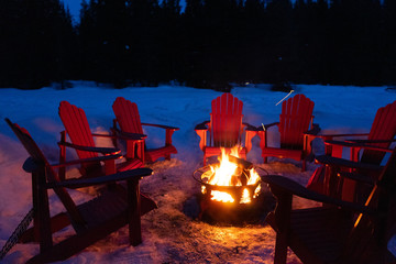 Cozy/romantic evening scene of warming bonfire on snow in winter with red chairs around. Canadian...