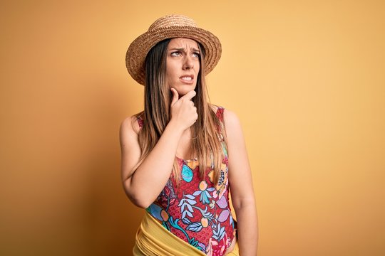 Young beautiful blonde woman wearing swimsuit and summer hat over yellow background Thinking worried about a question, concerned and nervous with hand on chin