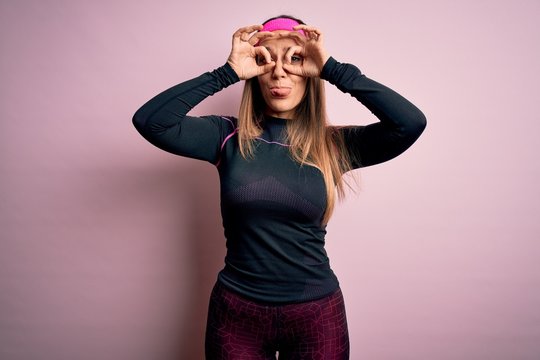 Young blonde fitness woman wearing sport workout clothes over isolated background doing ok gesture like binoculars sticking tongue out, eyes looking through fingers. Crazy expression.