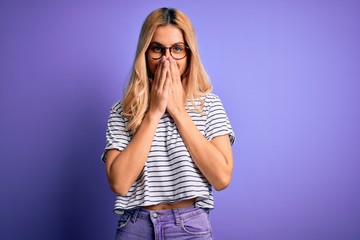 Young beautiful blonde woman wearing striped t-shirt and glasses over purple background laughing and embarrassed giggle covering mouth with hands, gossip and scandal concept