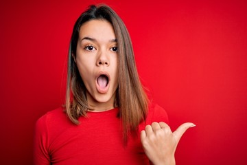 Young beautiful brunette girl wearing casual t-shirt over isolated red background Surprised pointing with hand finger to the side, open mouth amazed expression.