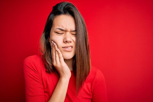 Young beautiful brunette girl wearing casual t-shirt over isolated red background touching mouth with hand with painful expression because of toothache or dental illness on teeth. Dentist concept.