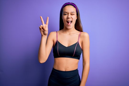 Young beautiful sporty girl doing sport wearing sportswear over isolated purple background smiling with happy face winking at the camera doing victory sign with fingers. Number two.