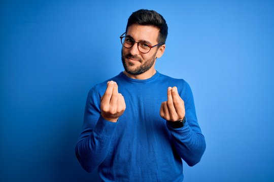 Young handsome man with beard wearing casual sweater and glasses over blue background doing money gesture with hands, asking for salary payment, millionaire business