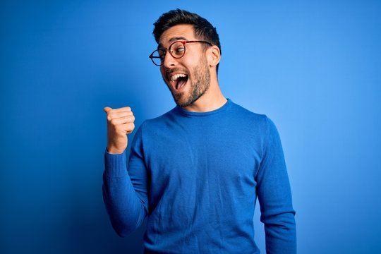 Young handsome man with beard wearing casual sweater and glasses over blue background smiling with happy face looking and pointing to the side with thumb up.