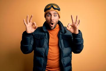 Young handsome skier man with beard wearing snow sportswear and ski goggles looking surprised and shocked doing ok approval symbol with fingers. Crazy expression