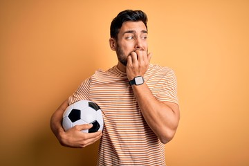 Handsome player man with beard playing soccer holding footballl ball over yellow background looking stressed and nervous with hands on mouth biting nails. Anxiety problem.