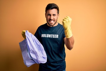 Young handsome volunteer man with beard cleaning junk using bag over yellow background annoyed and frustrated shouting with anger, crazy and yelling with raised hand, anger concept
