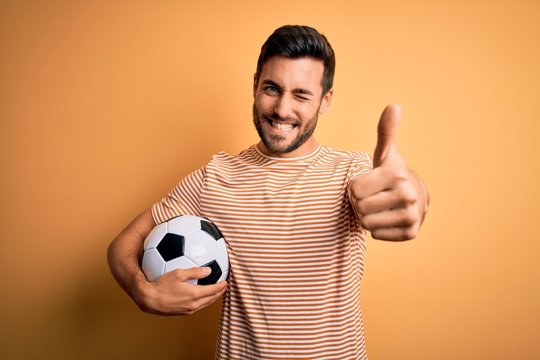 Fototapeta Handsome player man with beard playing soccer holding footballl ball over yellow background approving doing positive gesture with hand, thumbs up smiling and happy for success. Winner gesture.
