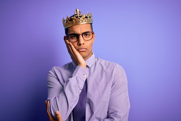 Young handsome african american man wearing golden crown of king over purple background thinking looking tired and bored with depression problems with crossed arms.