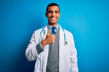 Handsome african american doctor man wearing coat and stethoscope over blue background doing happy thumbs up gesture with hand. Approving expression looking at the camera showing success.