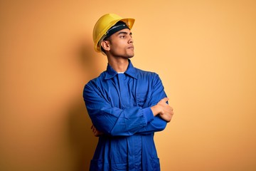 Young handsome african american worker man wearing blue uniform and security helmet looking to the side with arms crossed convinced and confident - Powered by Adobe
