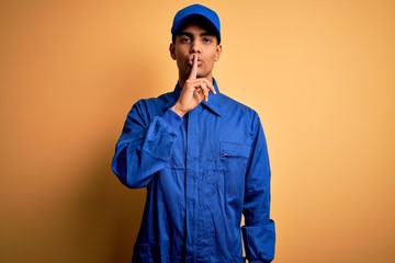 Young african american mechanic man wearing blue uniform and cap over yellow background asking to be quiet with finger on lips. Silence and secret concept.