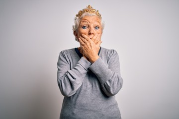 Senior beautiful grey-haired woman wearing golden queen crown over white background shocked covering mouth with hands for mistake. Secret concept.