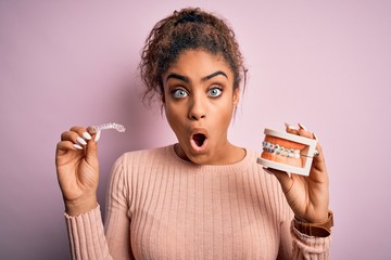 Young african american woman surprised and shocked holding professional orthodontic denture with...