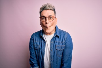 Young handsome modern man wearing glasses and denim jacket over pink isolated background making fish face with lips, crazy and comical gesture. Funny expression.