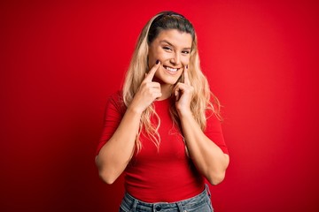 Fototapeta premium Young beautiful blonde woman wearing casual t-shirt standing over isolated red background Smiling with open mouth, fingers pointing and forcing cheerful smile