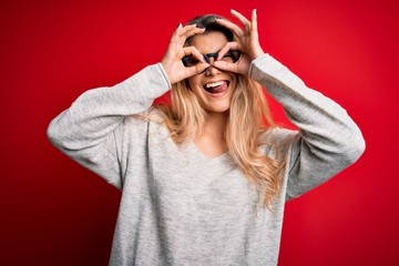 Young beautiful blonde woman wearing sweater and glasses over isolated red background doing ok gesture like binoculars sticking tongue out, eyes looking through fingers. Crazy expression.