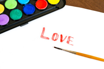 Handwritten LOVE text with brush and watercolor paints on white background. Box of watercolor paints with a brush for drawing. Love concept