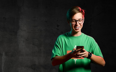 Young positive teen boy in green t-shirt and glasses standing, texting message on phone and smiling over dark background