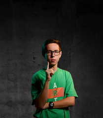 Young teen boy in green t-shirt, jeans and glasses standing, holding head, screaming and feeling shock over dark background