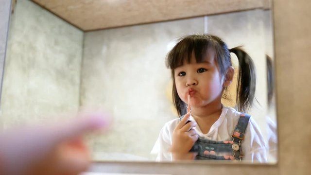 Cute child little girl doing makeup and having fun applying lipstick at a mirror in room.