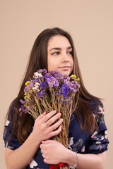 Portrait of a girl in the studio on a beige background. Dressed in a light blue dress. Presses a bouquet of wildflowers to his chest. Looking away. Vertical photo