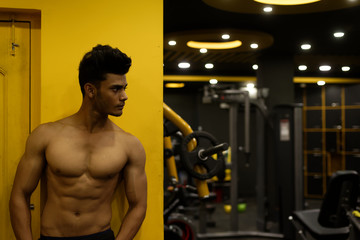 Fototapeta na wymiar Portrait of an young and handsome Indian Bengali brunette man with muscular body standing in a vibrant yellow background in a multi gym. Fitness and Indian lifestyle.