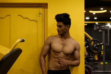 Fototapeta na wymiar Portrait of an young and handsome Indian Bengali brunette man with muscular body standing in a vibrant yellow background in a multi gym. Fitness and Indian lifestyle.