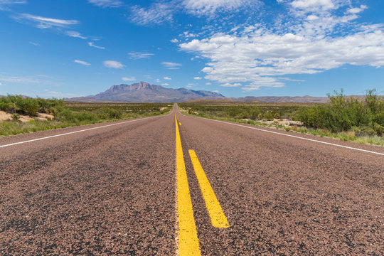 Long straight road in the desert, leading to a beautiful mountain range under a blue sky with clouds