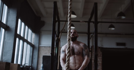 Fototapeta na wymiar Shirtless young athletic Caucasian sportsman climbing rope during extreme functional physical workout in large gym.